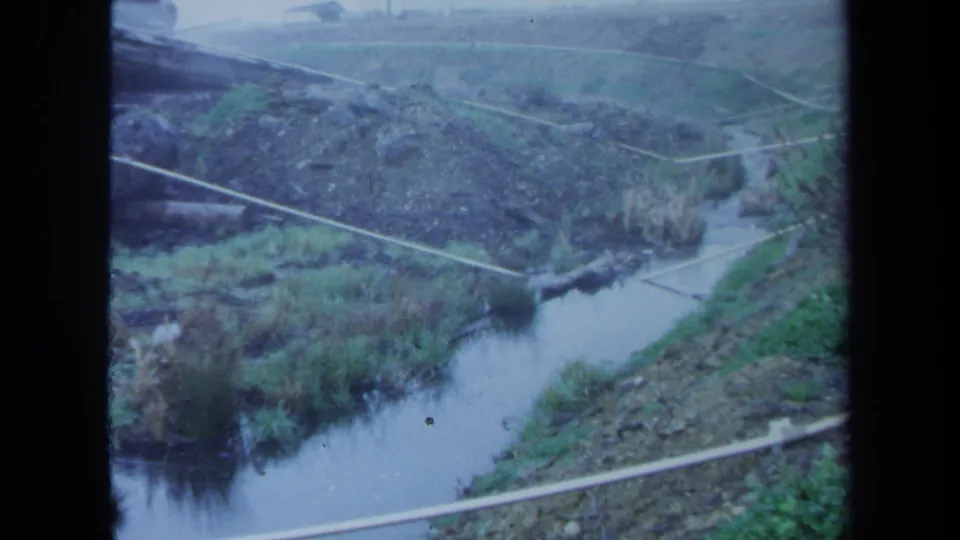 1971 Maine 8mm still showing an overhead view of a stream channel, pipe crossings, muddy banks, and rough rural ground.