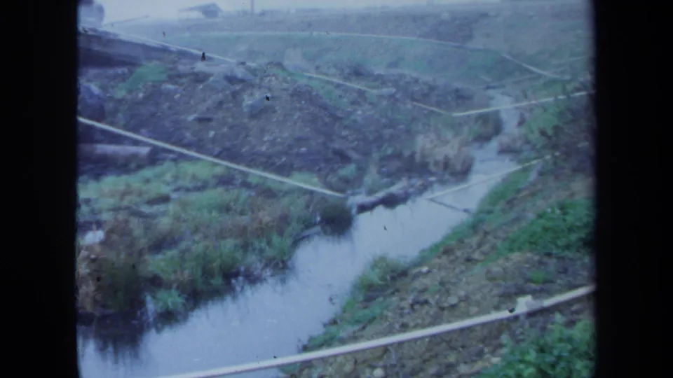 Faded 8mm frame from 1971 Maine showing pale pipes over a small stream in an overgrown rural or undeveloped setting.