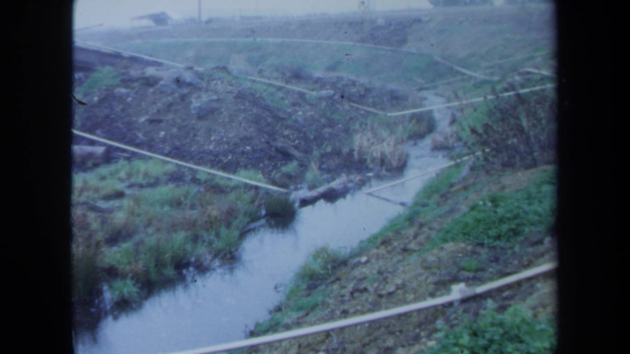 Narrow river winding through grassy embankments with brush and pale paths or rails crossing the slope.