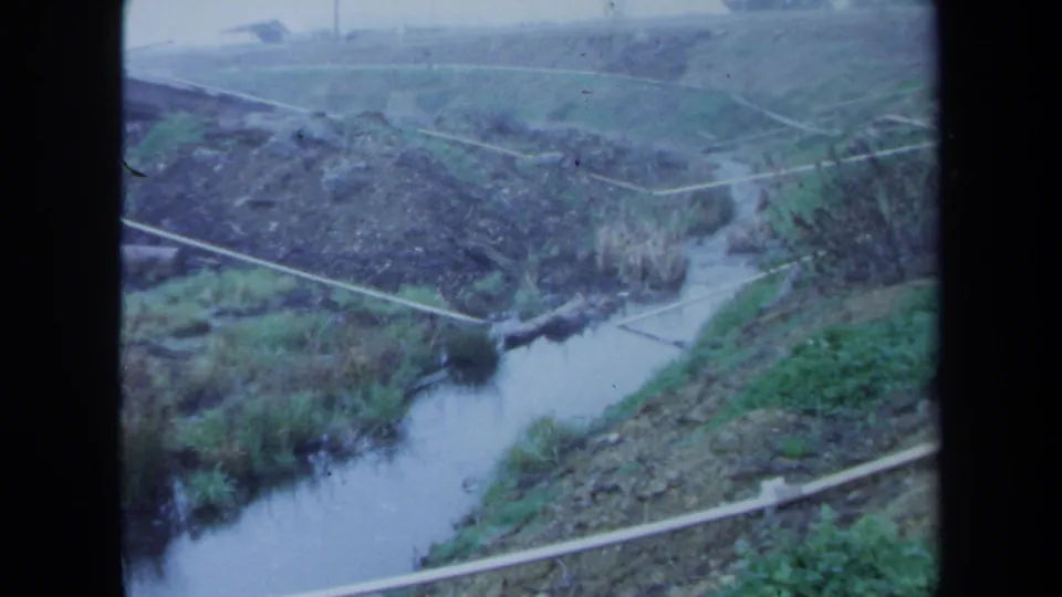 1971 Maine 8mm view of a small river below grassy, uneven embankments with brush and pale lines across the hillside.