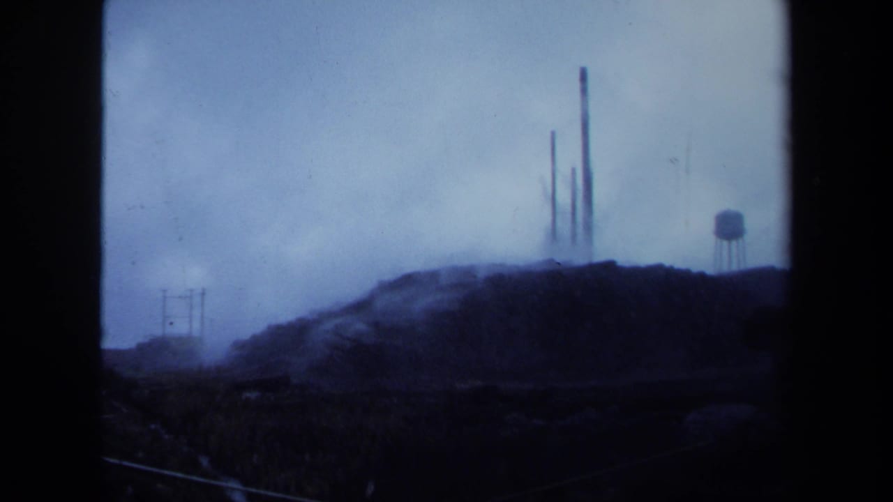 Distant smokestacks and a water tower rise beyond a dark hillside under foggy blue light.