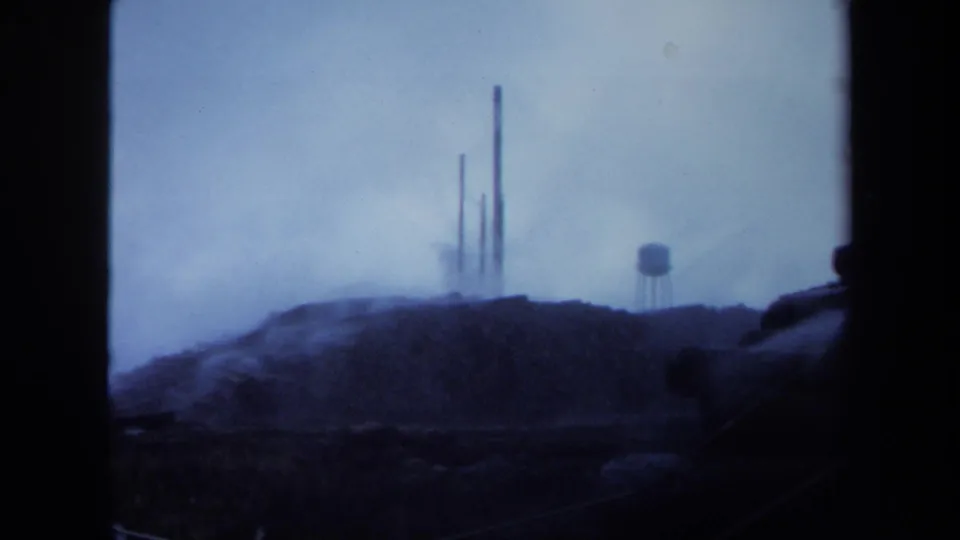 1971 Maine 8mm view of tall smokestacks and a water tower seen through blue haze, with a dark foreground ridge below.
