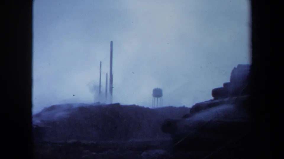 1971 Maine 8mm frame of smokestacks and a water tower in foggy distance, with dark hillside and film gate shadows along the edges.