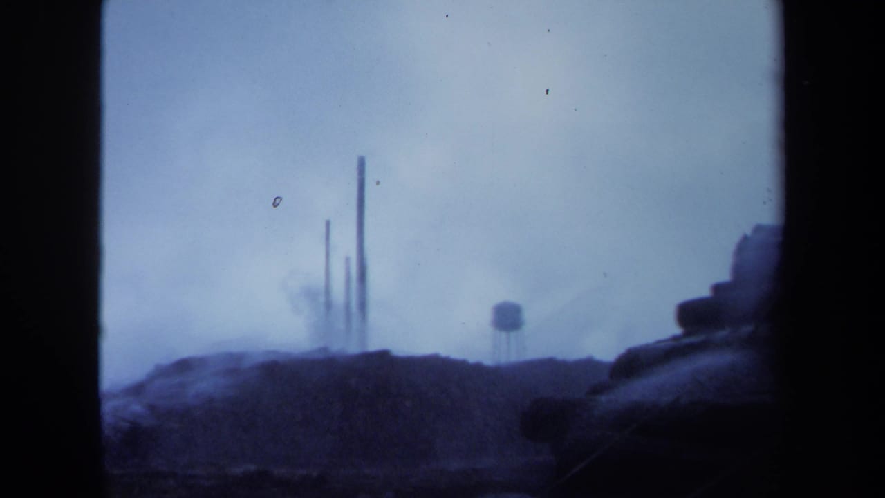 1971 Maine 8mm frame showing distant smokestacks, drifting smoke, low terrain, and a small water tower through dark borders.