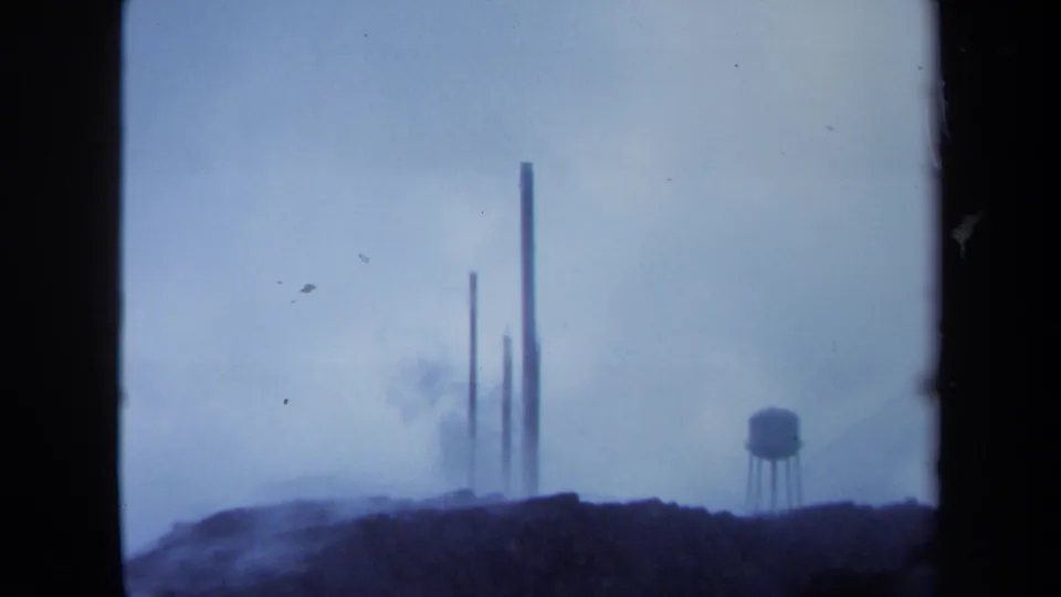 1971 Maine 8mm frame with several distant smokestacks, smoke haze, and a water tower at right in a soft blue cast.