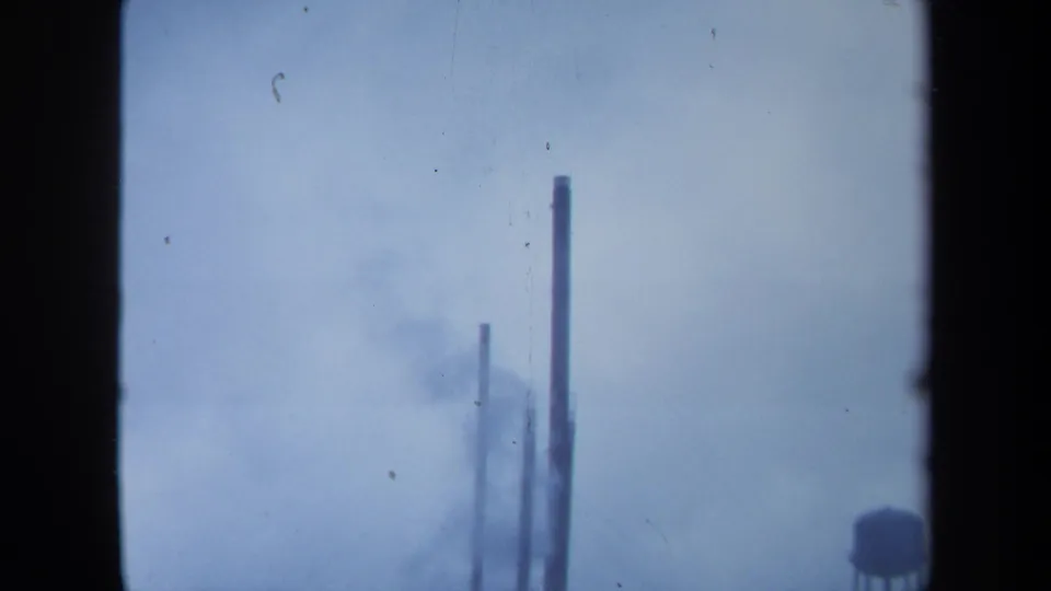 1971 Maine 8mm frame where tall stacks rise into haze, with the water tower partly visible at lower right and heavy film borders.
