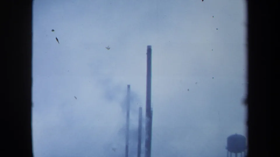 1971 Maine 8mm frame showing smoke drifting around distant stacks, a water tower near the right edge, and visible dust marks.
