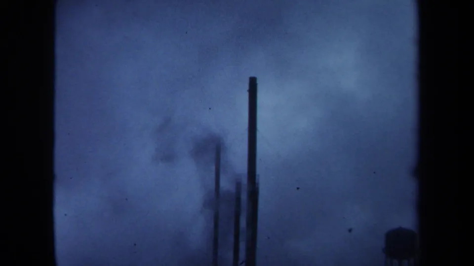 Blue-shifted 1971 Maine 8mm view of industrial stacks and smoke haze near a distant water tower.