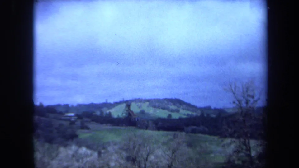 1971 Maine 8mm landscape view with green hills, sparse trees, and distant buildings below a cloudy sky.