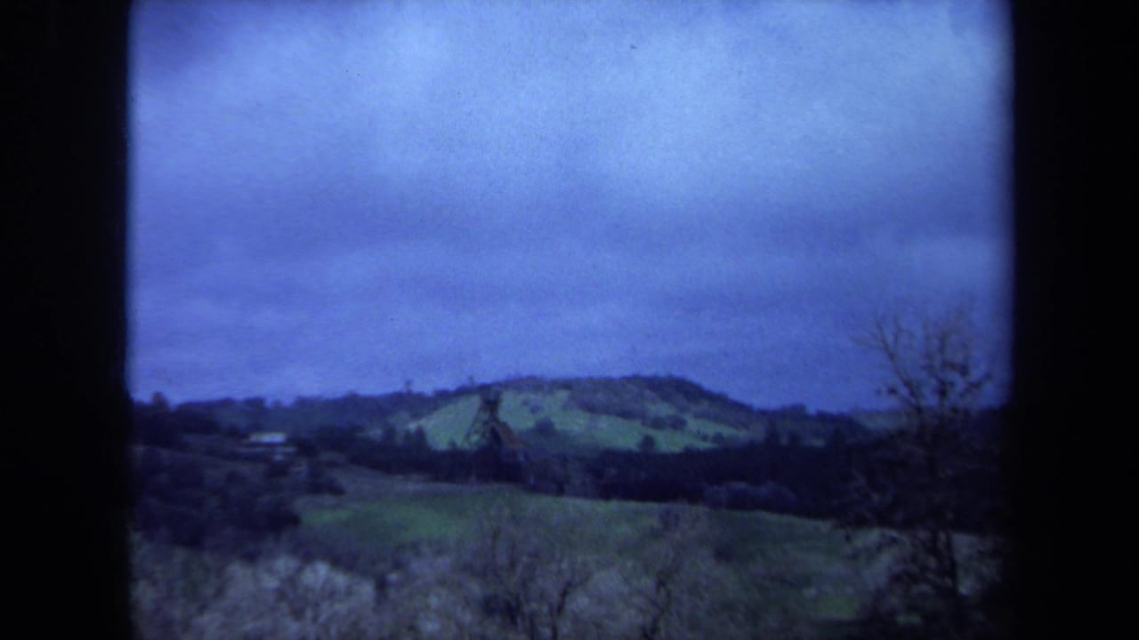 1971 Maine 8mm frame showing rolling green fields, a dark tree line, and a distant brown tower beneath a blue overcast sky.