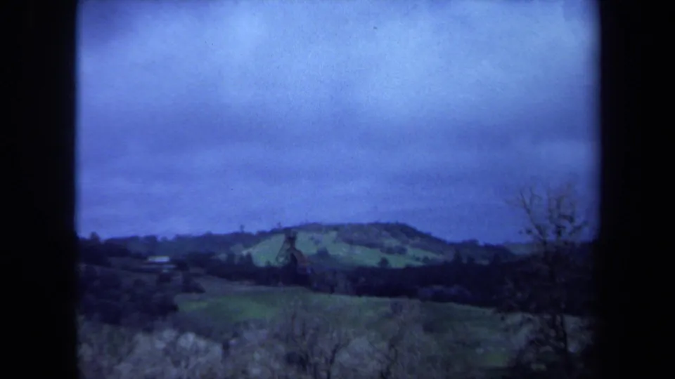 1971 Maine 8mm frame showing rolling green fields, a dark tree line, and a distant brown tower beneath a blue overcast sky.