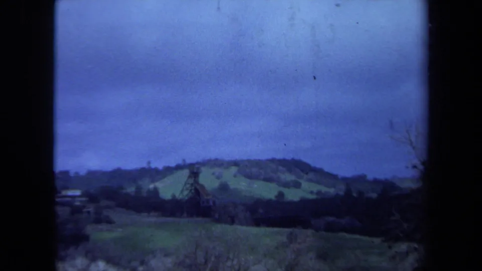 1971 Maine 8mm view of a brown tower set against green hills, dark trees, and a muted blue sky.