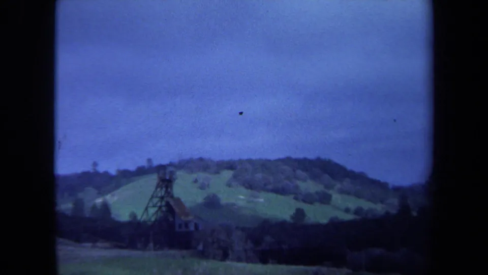 1971 Maine 8mm frame with the tower larger in view, backed by grassy hills, scattered trees, and a cloudy blue sky.