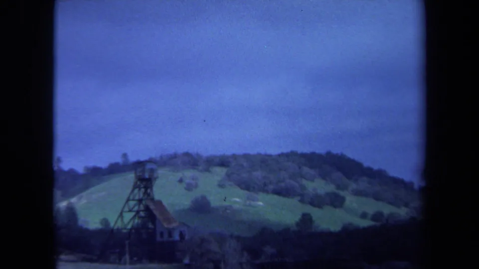 1971 Maine home-movie view of a rural tower structure set below wooded hills and a blue overcast sky.