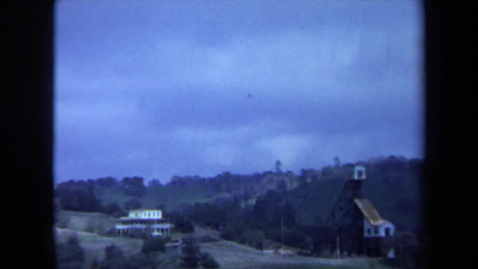 1971 Maine 8mm frame with a white house, rolling fields, trees, and a dark tower structure in the distance.