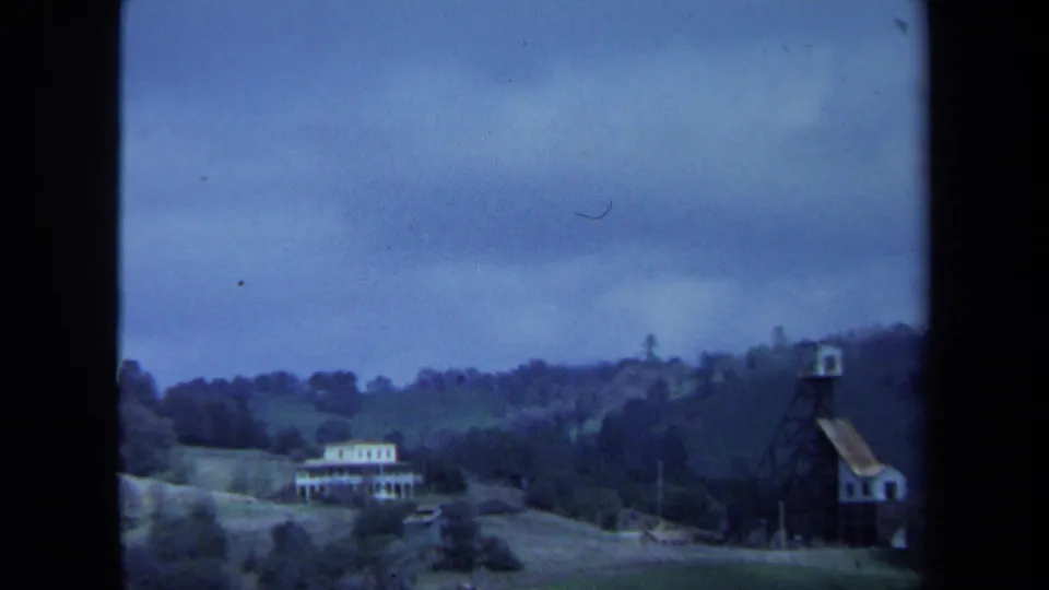 1971 Maine rural view showing a white house, open fields, wooded hills, and the tower structure near the frame edge.