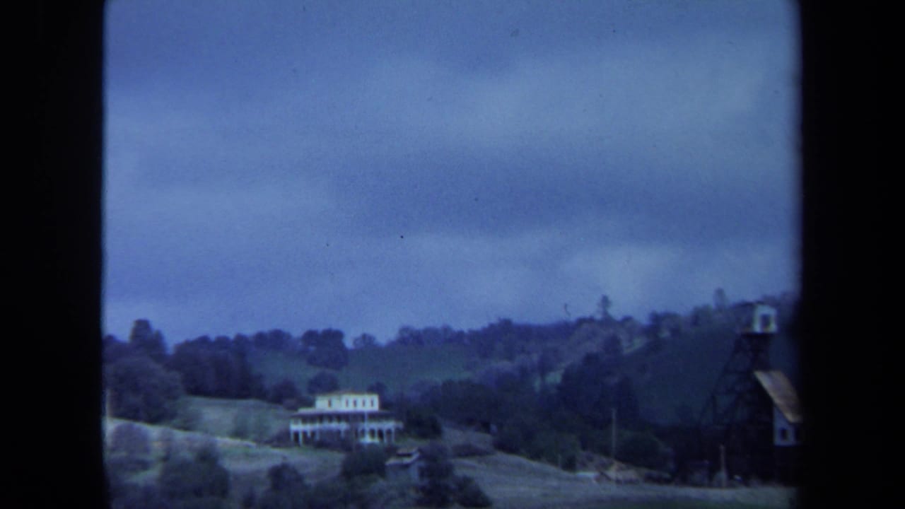 1971 Maine 8mm frame showing a distant white hillside house with porches, trees, rolling land, and a blue-shifted overcast sky.