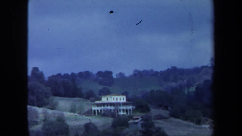 1971 Maine home-movie frame of a white house on a slope, surrounded by trees and rolling hills under a muted blue sky.
