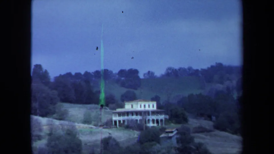 1971 Maine 8mm frame with the white hillside house, surrounding trees, rolling terrain, and a visible green film artifact near the left side.