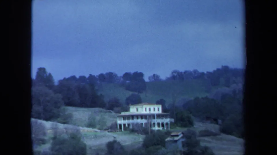 1971 Maine 8mm frame holding on a white porch-fronted house set into a grassy hillside with dense trees beyond.