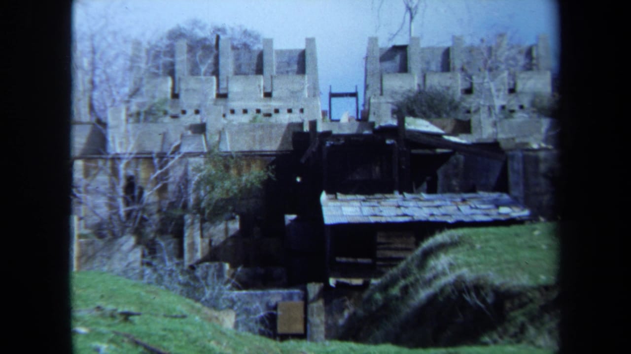 Stone battlement-like ruins, bare trees, grass, and a dark shed seen from across a slope.