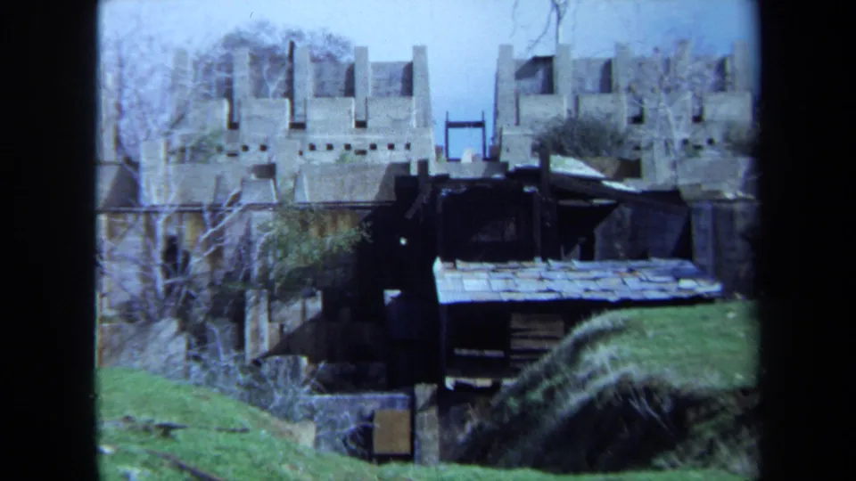 1971 Maine 8mm frame showing weathered stone remains, a small shed, green grass, and bare trees from a home-movie viewpoint.