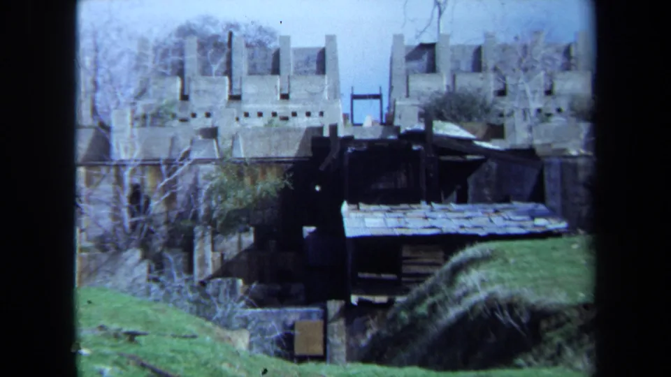 1971 Maine 8mm frame of castle-like ruins with a dark shed in front, green grass at right, and bare trees near the stonework.