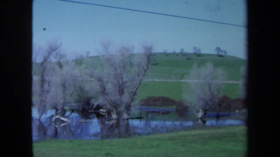1971 Maine home-movie frame with bare trees in floodwater, a grassy bank, hilltop trees, and visible 8mm gate borders.