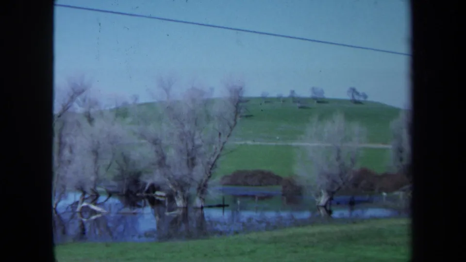 1971 Maine 8mm frame of a quiet flooded field, bare trees, grassy slope, pale sky, and a diagonal overhead line.