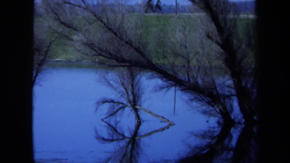 1971 Maine 8mm frame of a quiet waterside landscape with bare trees, blue water, grassy banks, and a distant utility line.