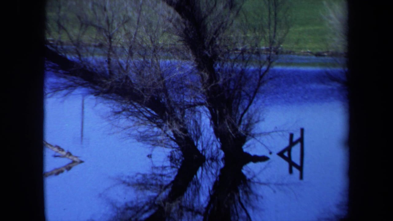 1971 Maine 8mm frame of still blue water reflecting bare trees and shoreline, with a small wooden A-frame shape near the right side.