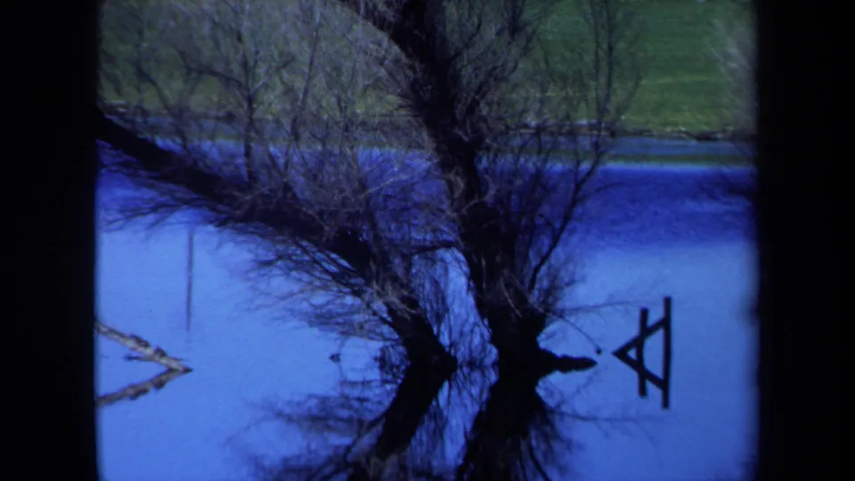 1971 Maine 8mm frame with bare shoreline trees mirrored in blue water and a small wooden A-frame shape near the right edge.