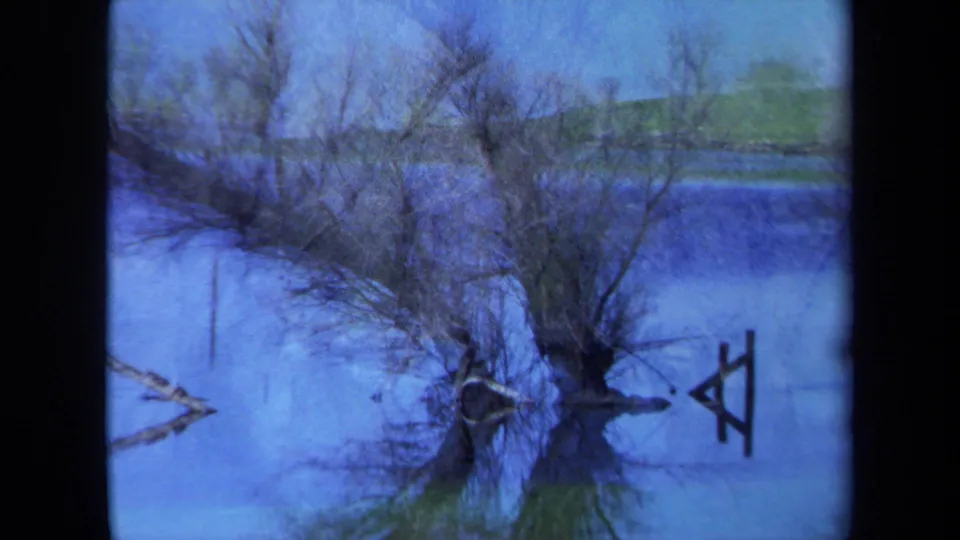 1971 Maine 8mm frame showing a softly exposed lakeside reflection, with bare trees, blue water, and a small wooden form.
