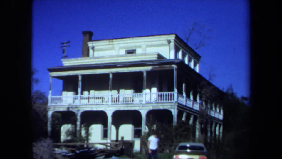 1971 Maine 8mm view of a large waterfront-style house with wraparound porch, parked car, and a distant person walking nearby.