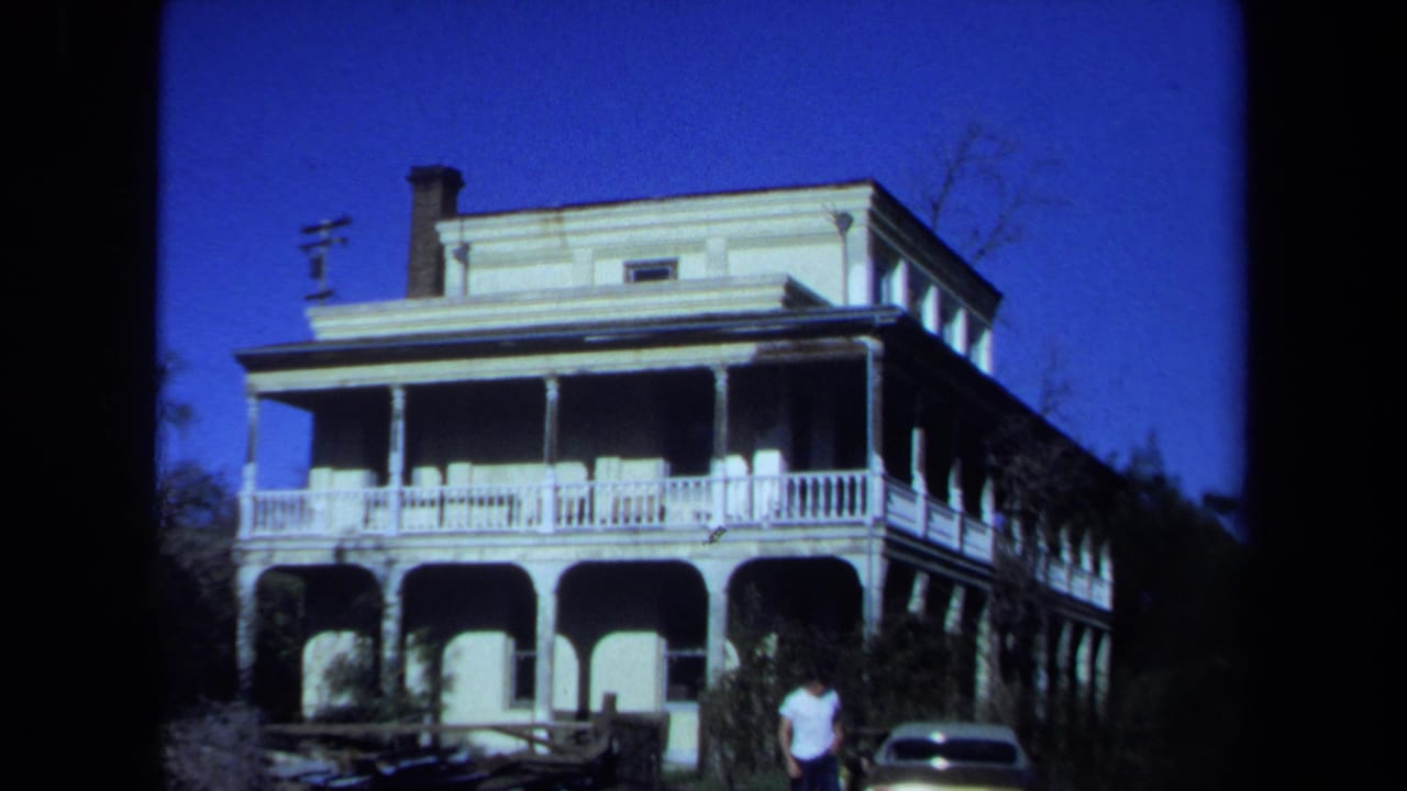 1971 Maine 8mm frame showing a large porch-fronted house with arched lower openings, white railings, chimney, trees, and a person near the entrance.