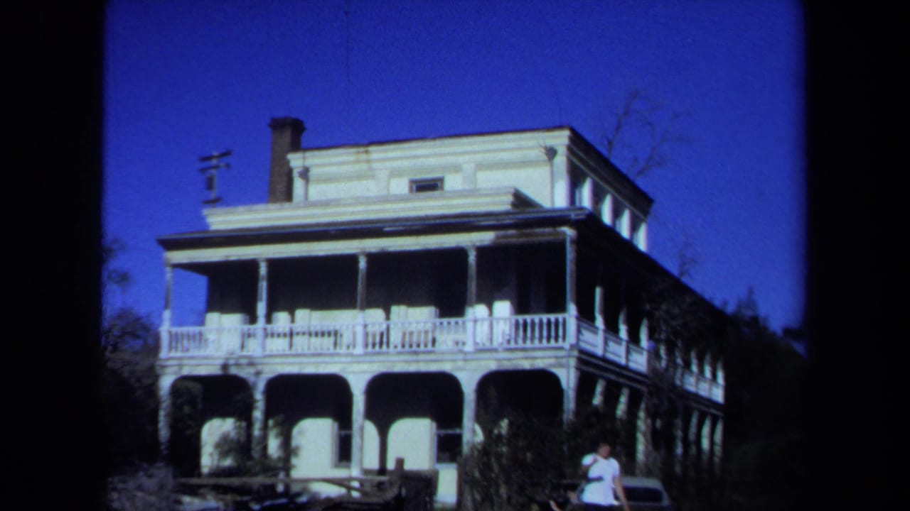 1971 Maine home-movie frame with a young man in a white T-shirt walking near a parked car in front of a large arched-porch house.