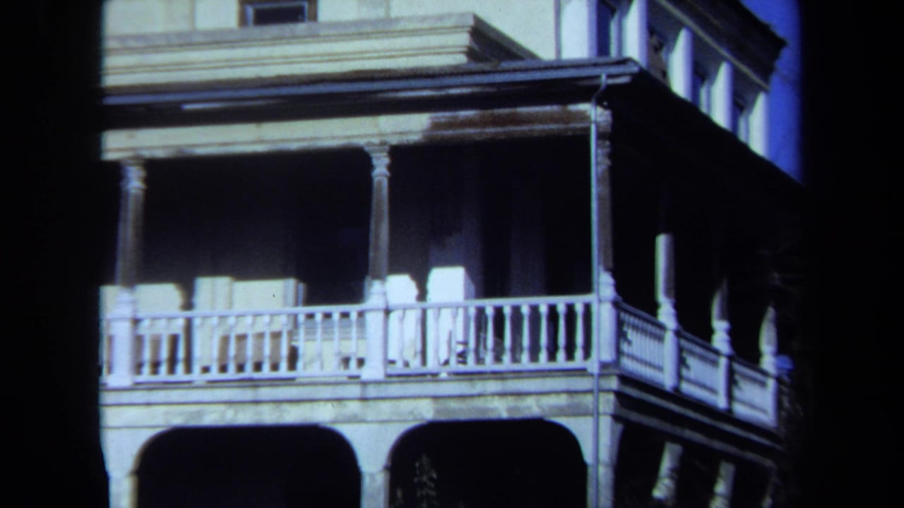 Weathered multi-story house with a wraparound porch and white railings under blue sky.