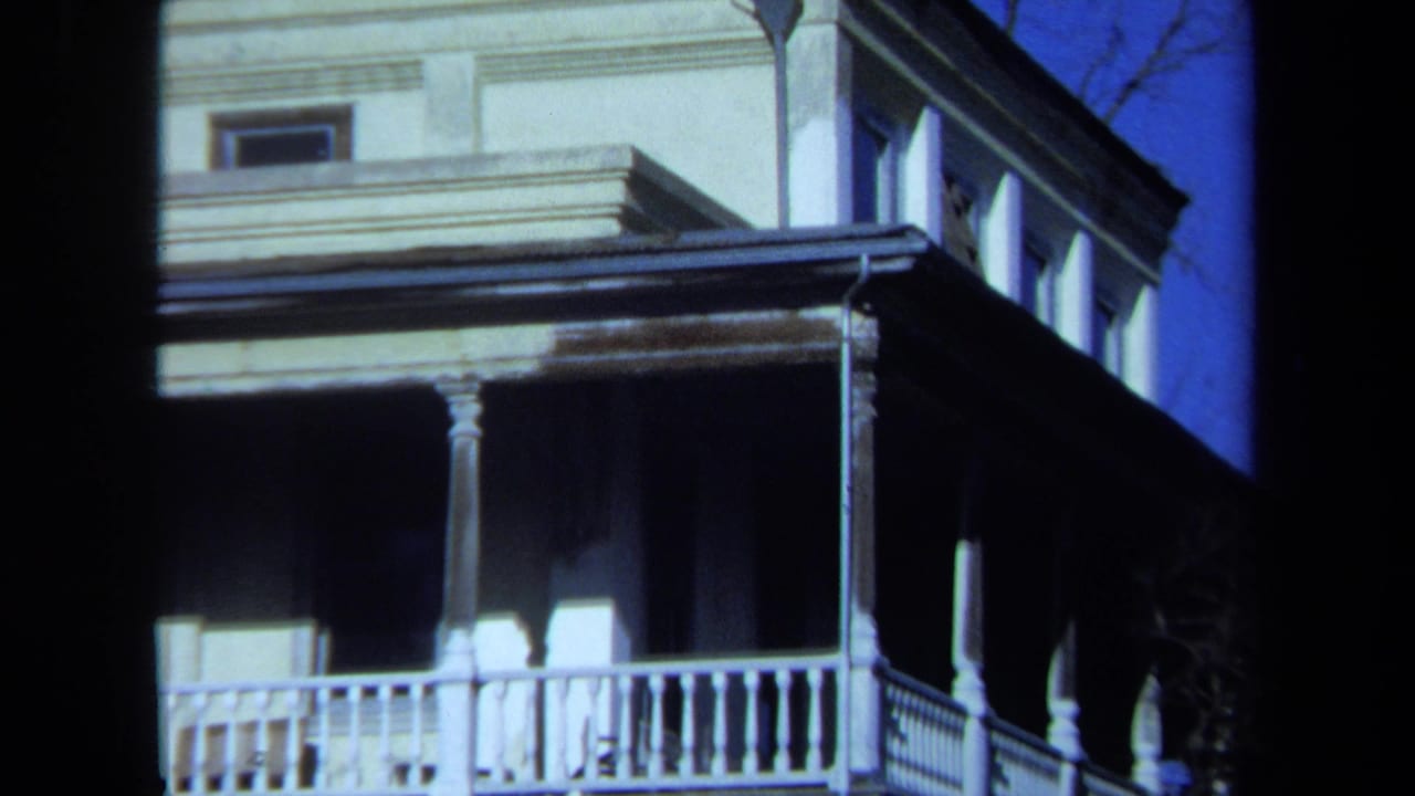 1971 Maine home-movie frame of an aging house facade, emphasizing the porch roof, white railing, columns, upper windows, and washed paint.