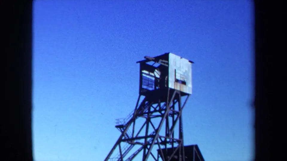 1971 Maine 8mm frame showing a dark metal industrial tower with a small enclosed top section against a bright blue sky.