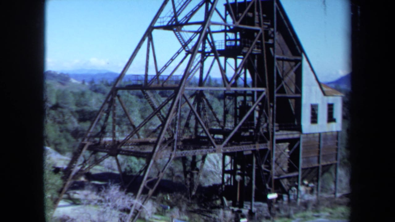 1971 Maine 8mm frame showing an aging metal industrial framework, attached building, trees, and distant hills within dark film borders.