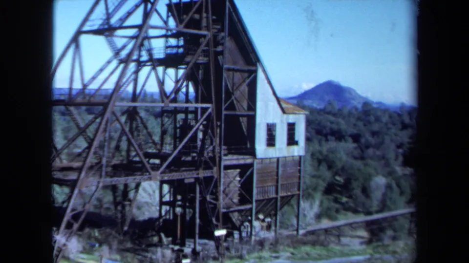 1971 Maine home-movie frame with a steel mill or mine-like structure, small attached building, forest, and distant mountain landscape.