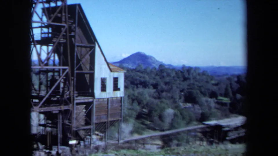 1971 Maine 8mm frame during a pan past the industrial building, revealing more forest, mountain slope, and a low conveyor-like structure.