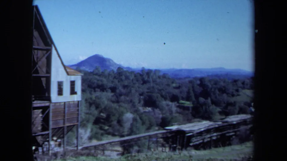 1971 Maine home-movie frame showing the edge of an old industrial building, wooded hills, a mountain, and a low track or conveyor in the foreground.