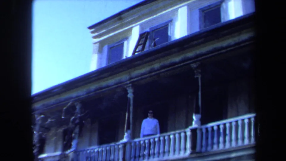 1971 Maine 8mm frame showing a woman standing on a white porch with railings, columns, windows, and shaded siding.