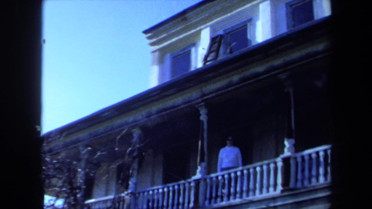 1971 Maine 8mm frame showing a man on a weathered house balcony with white railing, upper windows, and bare vines.