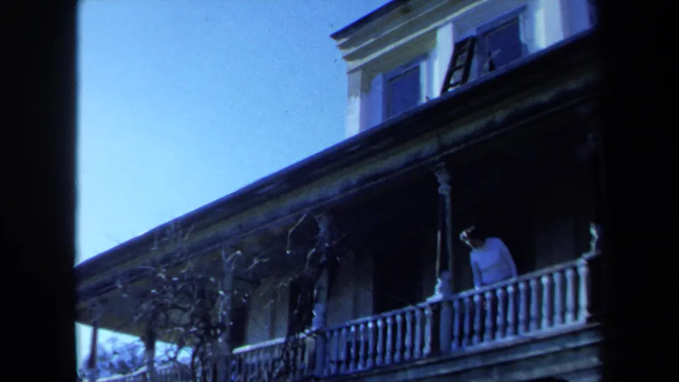 1971 Maine 8mm frame showing the man bending or peering over the white balcony railing on a weathered house exterior.