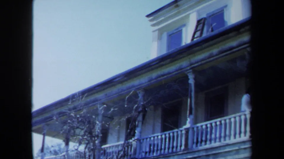 1971 Maine home-movie frame with the man near the right edge of the balcony, a white railing, porch roofline, vines, and upper window.