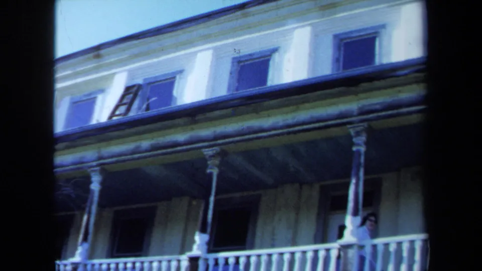 1971 Maine home-movie frame with a person on the upper porch of a weathered mansion with columns and railings.
