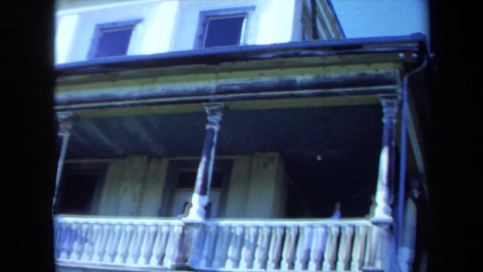 1971 Maine 8mm frame showing a close view of the mansion porch, with railings, columns, shaded doorway, and aged siding.