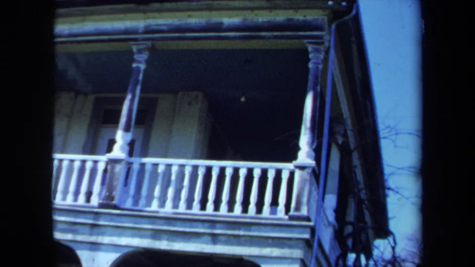 1971 Maine 8mm frame showing the corner of a weathered mansion porch with white columns, railings, and shaded exterior wall.